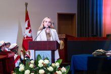 Ministra Zdrowia stoi za mównicą i mówi do mikrofonu. W tle widać sztandar WUM. Po lewej stronie siedzą osoby w białych czapkach z czerwonymi paskami. Przed mównicą leży bukiet białych róż z zielonymi liśćmi. Po prawej stronie stoi stół z niebieskim obrusem, na którym leżą czapki akademickie.