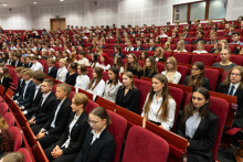 W dużej sali wykładowej z czerwonymi fotelami siedzi kilkadziesiąt osób. Większość to młode kobiety i mężczyźni w białych koszulach i ciemnych marynarkach. Wszyscy patrzą w stronę mównicy poza kadrem. Osoby siedzące zachowują skupienie. 