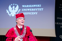 Mężczyzna w czerwonej todze i birecie, prorektor ds. studenckich i kształcenia, stoi przy mównicy i przemawia. W tle widać godło WUM w kolorze białym.