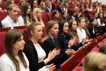 Immatrykulowani studenci, formalnie ubrani, siedzą w auli na czerwonych krzesłach i biją brawo. Patrzą przed siebie.