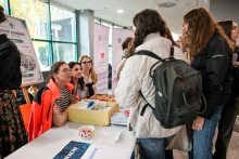 Kilka osób stoi przy stole w jasnym korytarzu. Na stole leży żółte pudełko z materiałami i miska w biało-czerwony wzór. Za stołem siedzą dwie osoby w pomarańczowych kurtkach i rozmawiają z odwiedzającymi. W tle widać plakaty informacyjne.