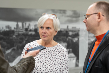 Kobieta z krótkimi białymi włosami i okularami na głowie, ubrana w białą koszulę w czarne i czerwone kropki, stoi obok mężczyzny w okularach, ubranym w pomarańczową koszulkę i ciemną marynarkę. Mówią do dyktafonu.