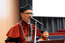 Dziekan wydziału, ubrany w czarnopomarańczową togę stoi przy mównicy i przemawia. 