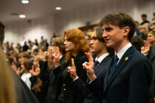 Grupa młodych osób, studentów pierwszego roku, stoi i wygłasza przysięgę studencką. Mają podniesione prawe dłonie i wyciągnięte z niej ku górze dwa palce.