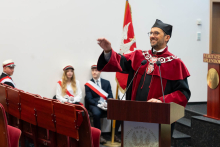 Mężczyzna w todze i czarnym birecie przemawia z mównicy, gestykuluje prawą ręką i uśmiecha się. Na mównicy widnieje herb Warszawskiego Uniwersytetu Medycznego. W tle widać flagę z orłem oraz cztery osoby w białych mundurach z czerwonymi szarfami, siedzące na krzesłach. Po prawej stronie znajduje się fragment napisu „VERITATIS SPLENDOR”.