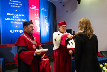 Immatrykulacja studentki. Dwóch mężczyzn w czerwonych togach i biretach stoi na scenie. Jeden trzyma berło, którym dotyka ramienia stojącej przed nim studentki.