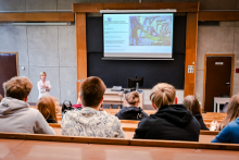 W sali wykładowej siedzi osiem osób w drewnianych ławkach. Patrzą na ekran z prezentacją. Przed ekranem stoi kobieta w jasnym stroju, która coś tłumaczy.
