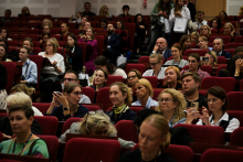 Duża grupa kobiet i mężczyzn siedzi w audytorium na czerwonych fotelach ustawionych w rzędach.