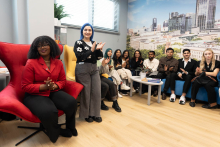 Grupa młodych osób, różnego pochodzenia, ubranych nieformalnie. Większość siedzi, jedna stoi. Na ścianie za nimi widać fototapetę kampusu WUM.