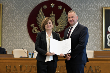 Dwie osoby, kobieta i mężczyzna, stoją obok siebie w formalnym otoczeniu. Osoba po lewej stronie trzyma otwarty dokument skierowany w stronę aparatu.