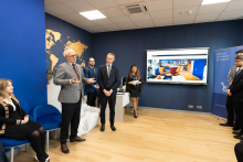 Grupa uczestników oficjalnego otwarcia. Część stoi, część siedzi. Po lewej stronie stoi rektor i przemawia.