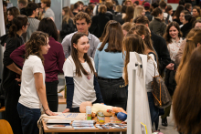 Kilka osób stoi przy stole w zatłoczonym korytarzu. Na stole leży fantom niemowlęcia w jasnym kolorze, kilka małych pojemników i ulotki. Dwie osoby w białych koszulkach rozmawiają z odwiedzającymi. W tle widać grupy ludzi przy innych stanowiskach.