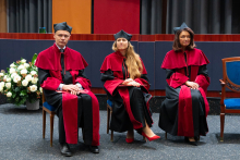 Trzy osoby: prodziekani, siedzą na krzesłach w sali. Noszą czarne togi z czerwonymi elementami i czarne birety. W tle widać zasłony i kolorowe panele ścienne. Obok nich stoi bukiet białych kwiatów.