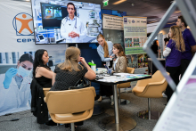 Na pierwszym planie widoczny jest stojak z broszurami. Widoczna broszura ma slogan „Odkryj z nami świat możliwości!” oraz zdjęcie miasta nocą. Nad nim wisi plakat z hasłem „TECHNOLOGIE INNOWACJE WSPÓŁPRACA”. W tle dwie osoby siedzą przy stole i rozmawiają. Po prawej stronie znajduje się stół z ulotkami. Dominują kolory biały, niebieski i czerwony.