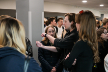 Kilka osób stoi w korytarzu. Jedna osoba w czarnej koszuli pokazuje coś dłonią, a druga w ciemnej kurtce patrzy na gest. Obok stoją osoby w jasnych i ciemnych ubraniach, część z plecakami. 