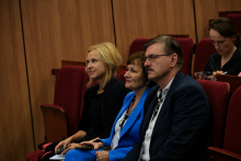 Dwie kobiety i mężczyzna siedzą w pierwszym rzędzie sali konferencyjnej lub audytoryjnej. Ubrane są formalnie, co sugeruje udział w oficjalnym wydarzeniu, takim jak konferencja lub wykład.