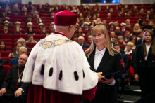 Rektor wręcza dyplom kobiecie w czarnym garniturze. W tle siedzi publiczność w akademickich togach i biretach. Sala jest pełna. 