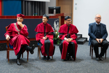 Cztery osoby siedzą na krzesłach w auli. Troje z nich, prodziekani i prorektor, osi czerwone togi i birety, jeden ma ciemny garnitur - to prof. Czupryniak. 