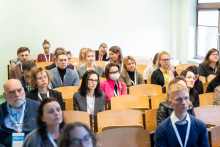 Grupa osób w strojach biznesowych siedzi w rzędach drewnianych krzeseł w sali wykładowej. Niektórzy mają identyfikatory na smyczach. Po prawej stronie znajduje się okno.