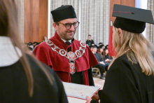 Dziekan w bordowej todze trzyma dokument. Naprzeciwko stoi osoba w czarnej todze i birecie, z długimi blond włosami, również trzyma dyplom. W tle siedzą osoby w czarnych togach.