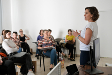 Grupa kobiet i mężczyzn siedzi w sali konferencyjnej, zwrócona w stronę kobiety prowadzącej prezentację. Prowadząca stoi obok biurka, na którym znajduje się monitor komputera wyświetlający slajdy. 