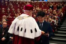 Rektor wręcza dyplom mężczyźnie w czarnym garniturze. W tle siedzi publiczność w akademickich togach i biretach. Sala jest pełna. 