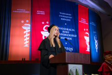 Młoda kobieta, przewodnicząca Samorządu Studentów, w czarnej todze i birecie stoi przy mównicy i przemawia. W tle wiszą kolorowe banery z nazwami wydziałów i napisem „Inauguracja Roku Akademickiego 2025/2026”. 