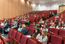 A lecture hall with red armchairs, in which a large group of young people are seated.
