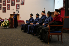 Pięść osób w togach i czarnych biretach siedzi na scenie. To prodziekani i zaproszony wykładowca. Dziekan Wydziału stoi przy mównicy i przemawia. W tle wiszą portrety. Na podłodze obok mównicy leży bukiet z białych kwiatów.