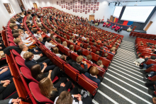 Tłum ludzi siedzących w auli. Większość patrzy na scenę, gdzie stoi grupa osób w czerwonych togach.