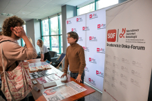 Kilka osób stoi przy stole w jasnym pomieszczeniu. Na stole leżą ulotki i plakaty z grafikami medycznymi. Jedna osoba w brązowym swetrze rozmawia z odwiedzającym w jasnej bluzie. W tle widać biały baner z czerwonym logo „SOF” i napisem „Studenckie Onko-Forum”