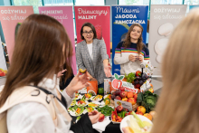 Grupa osób zgromadzona wokół stołu z talerzami pełnymi pokrojonych owoców i warzyw oraz całymi jabłkami, paprykami i liściastymi warzywami. 