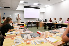 Kobieta w jasnej marynarce stoi w sali konferencyjnej i prowadzi prezentację, trzymając mikrofon. Przed nią znajduje się ekran projekcyjny z wyświetlonym slajdem zawierającym tekst. Wokół stołów ustawionych w kształcie litery „U” siedzi grupa kobiet i mężczyzn