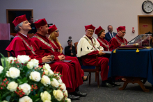 Sześć osób w czerwonych togach i biretach - prorektorzy, siedzący na środki auli w czasie uroczystości. Na pierwszym planie po lewej stronie stoi bukiet białych i czerwonych kwiatów. Scena przedstawia moment uroczystości akademickiej.