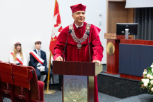 Prorektor w czerwonym stroju akademickim przemawia na podium z logo Warszawskiego Uniwersytetu Medycznego. Po jego prawej stronie stoi mównica z napisem „VERITATIS SPLENDOR”. W tle siedzą dwie osoby w galowych mundurach.