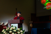 Kobieta w czarnej todze i birecie stoi przy mównicy ozdobionej herbem uczelni i bukietem białych róż. Nosi okulary. W tle widać ekran z napisem „Prof. dr hab. Magdalena Kucia” i obrazem komórek pod mikroskopem.