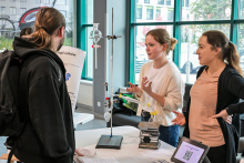 Trzy osoby stoją przy stole w jasnym pomieszczeniu z dużymi oknami. Na stole znajduje się zestaw do eksperymentów chemicznych z kolbami i statywem, obok leży tablet z kodem QR i kartki z informacjami. Jedna osoba w białej bluzie gestykuluje, druga w beżowej koszulce słucha, a trzecia w czarnej bluzie z plecakiem patrzy na stanowisko.