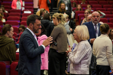 W centrum dwie osoby rozmawiają ze sobą – mężczyzna w garniturze i okularach oraz kobieta trzymająca dokumenty. W tle widoczne są inne osoby, siedzące lub stojące