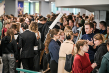 Duża grupa osób stoi w zatłoczonym korytarzu podczas wydarzenia. Ludzie rozmawiają w kilku mniejszych grupach, część trzyma ulotki. Widać osoby w ciemnych i jasnych ubraniach, w tym w garniturze i płaszczu.