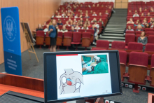 A lecture hall with an audience. Illustrations related to defibrillation are visible on a computer screen. The presenter is in the background.