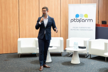 Mężczyzna w garniturze stoi na scenie z mikrofonem. W tle baner z logo i nazwą „ptbfarm” oraz tekstem „Polskie Towarzystwo Bezpiecznej Farmakoterapii”. Na scenie białe fotele i stoliki z butelkami wody.
