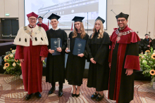 Pięć stojących osób. W środku trzy młode osoby ubrane w czarne togi. Dwie z nich trzymają w rękach granatowe teczki. Po lewej i po prawej - dwóch mężczyzn w średnim wieku, ubranych w bordowe togi. Wszyscy uśmiechają się do zdjęcia.