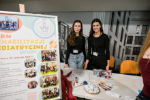 Dwie osoby stoją za stołem w jasnym pomieszczeniu. Na stole leżą ulotki, długopisy, identyfikatory i miska z losami. Obok stoi pluszowa maskotka w czerwonym kolorze. Po lewej stronie znajduje się duży plakat z napisem „SKN Rehabilitacji Pediatrycznej WUM” i zdjęciami grupowymi. W tle widać czarno-białą grafikę budynku i kilka krzeseł w kolorze pomarańczowym.