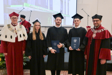 Pięć osób. Dwie osoby w środku mają czarne togi i birety, trzymają granatowe teczki z herbem WUM. Po bokach stoją dwie osoby w bordowych togach.