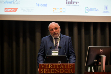 Kobieta z długimi, jasnymi włosami i okularami stoi za drewnianą mównicą podczas przemówieni