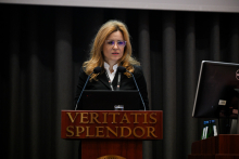 Kobieta z długimi, jasnymi włosami i okularami stoi za drewnianą mównicą podczas przemówienia