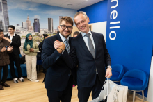 Prodziekan i dziekan uśmiechnięci pozują do zdjęcia. Prawe dłonie mają zwarte w uścisku. Za nimi widać innych uczestników oficjalnego otwarcia.