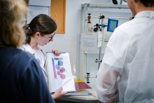 Trzy osoby stoją przy blacie w laboratorium. Jedna osoba trzyma otwarty segregator z kolorowymi ilustracjami roślin i podpisem „Barwy kwiatów”. Na blacie leżą dokumenty i czerwony folder. W tle widać tablicę korkową, sprzęt laboratoryjny.