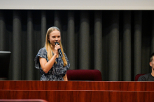 Młoda kobieta stoi za drewnianym pulpitem, trzyma mikrofon i przemawia. W tle widoczne są ciemne zasłony.