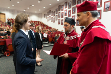 Młody mężczyzna w garniturze odbiera dyplom od dwóch profesorów w czerwonych togach i biretach. W tle siedzi liczna grupa studentów. 